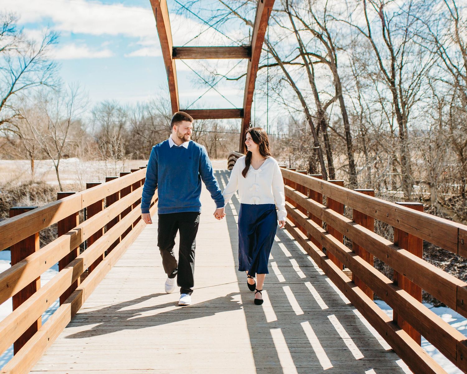 Happy engaged couple walking hand in hand on a sunny wooden bridge with bare winter trees behind.