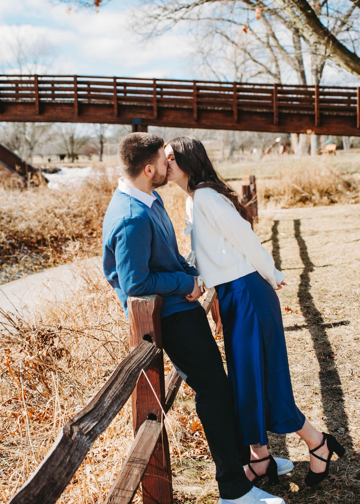 Couple sharing a romantic kiss near a wooden fence with a bridge in the background.