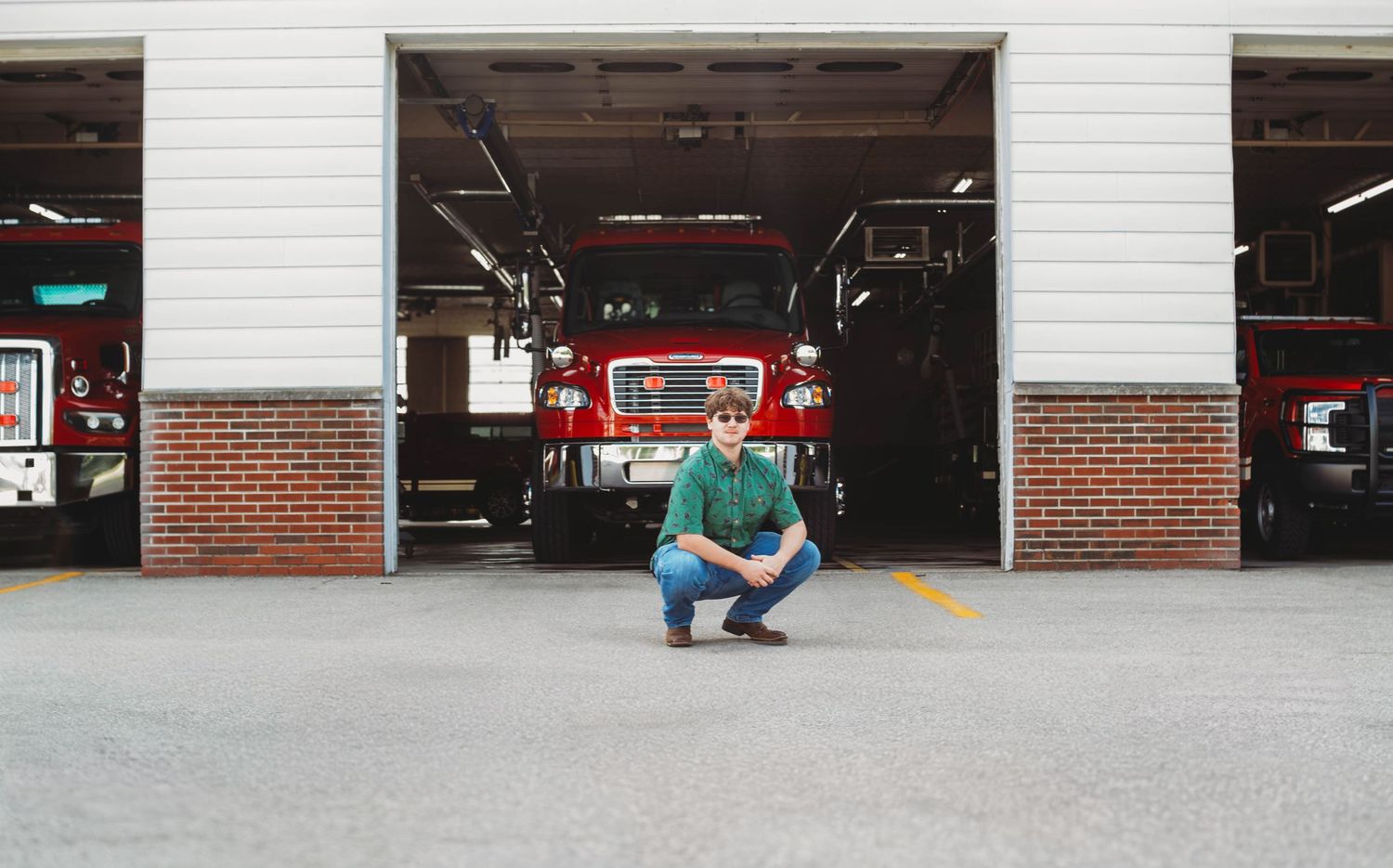 Young man crouching in front of a fire station with red fire trucks visible in the background.