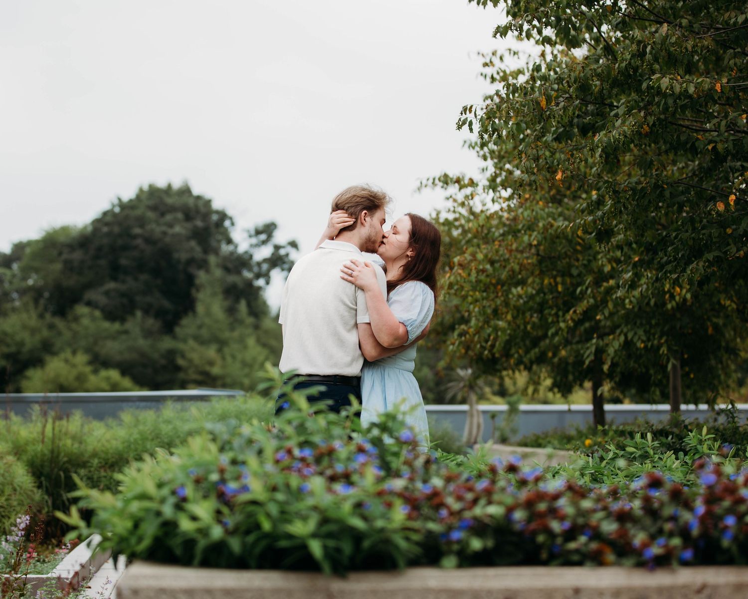 Couple sharing a romantic kiss surrounded by lush greenery and colorful flowers outdoors.