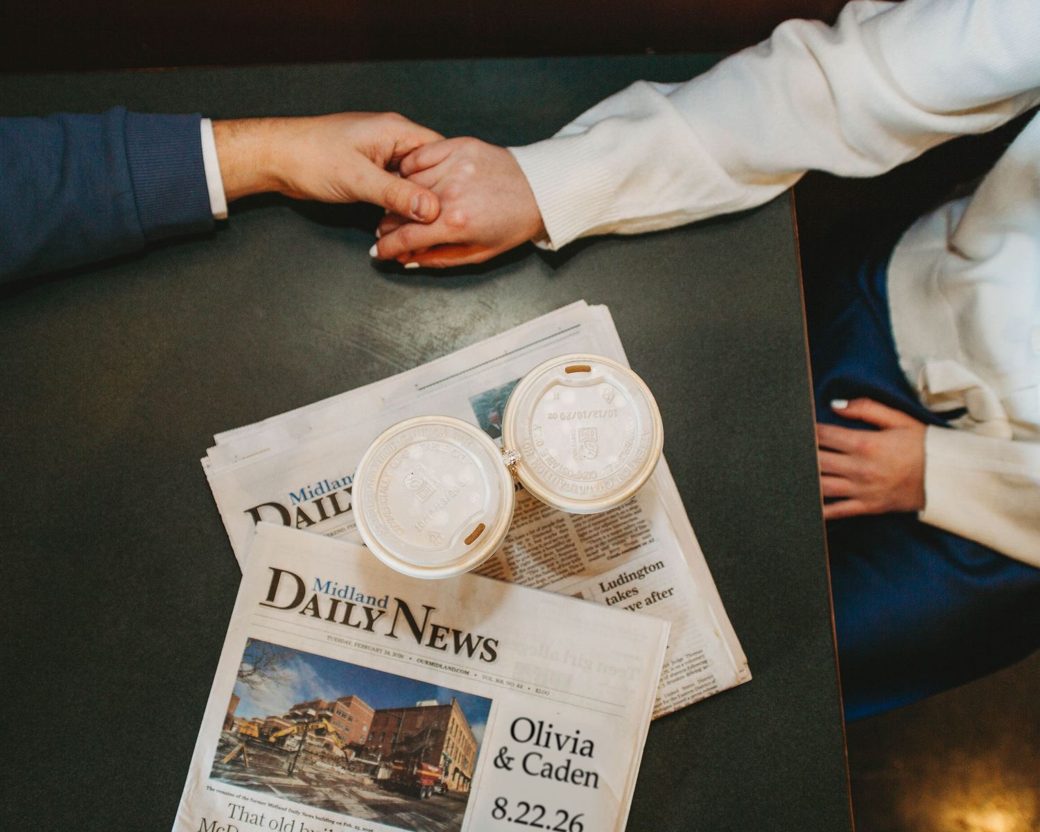 Overhead view of couple holding hands over a newspaper featuring their names Olivia and Caden 8.22.26.