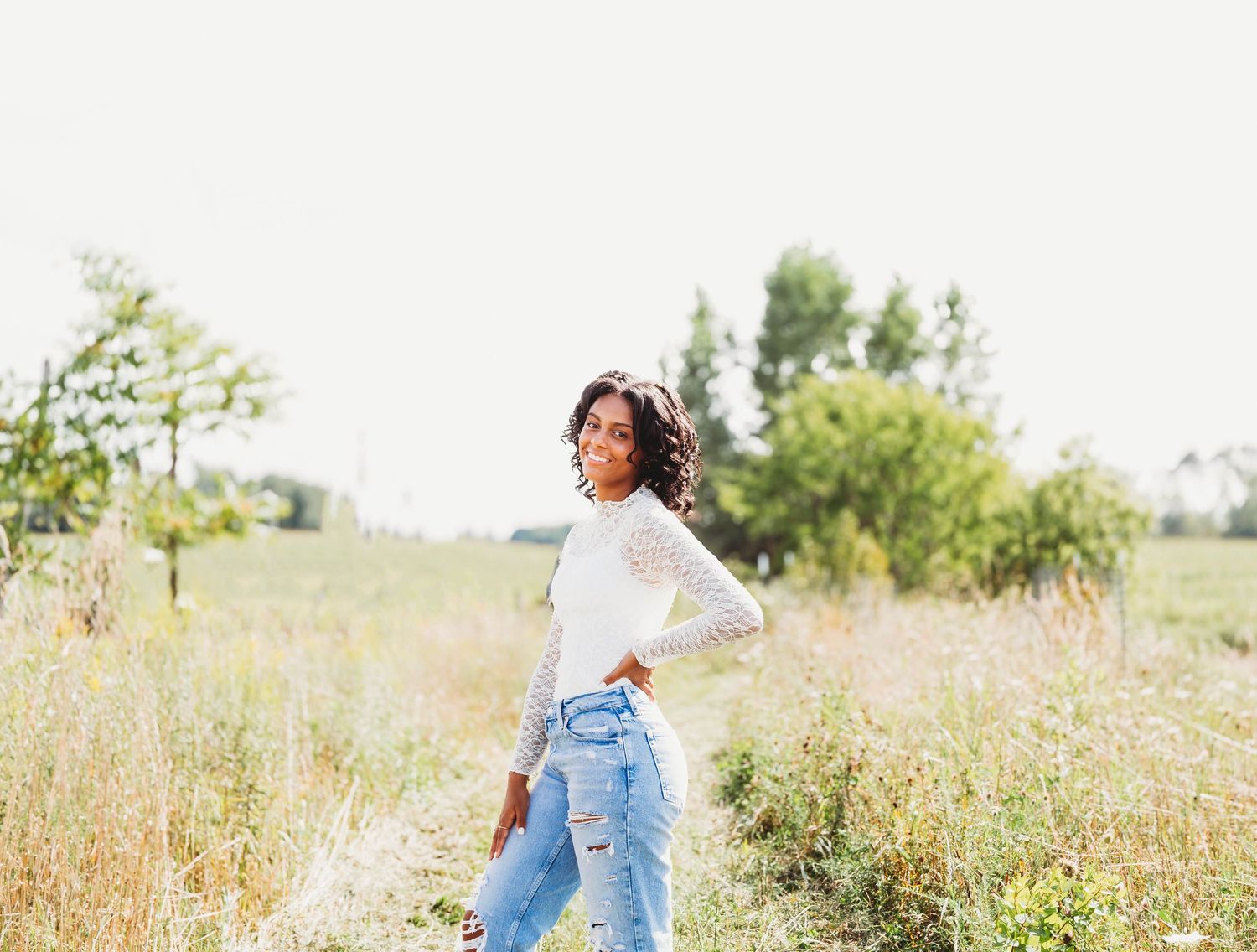 Young woman in white lace top and ripped jeans posing in a golden sunlit field outdoors.