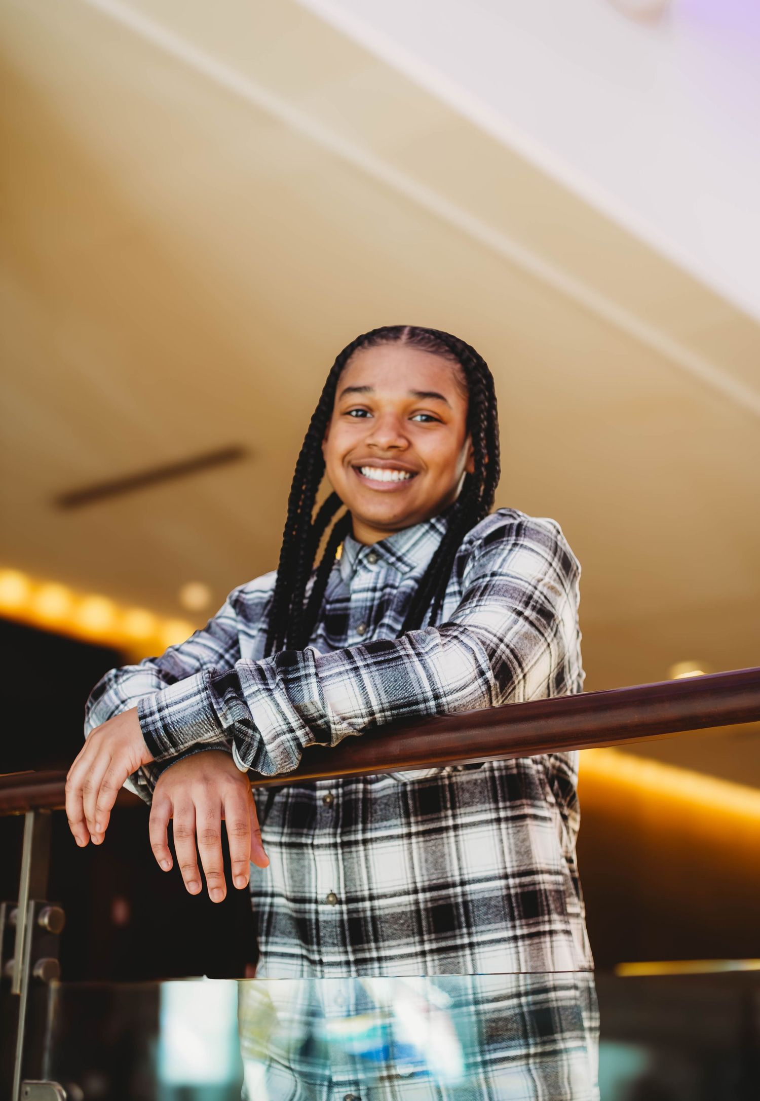 Smiling young woman with braids in plaid shirt leaning on railing indoors.