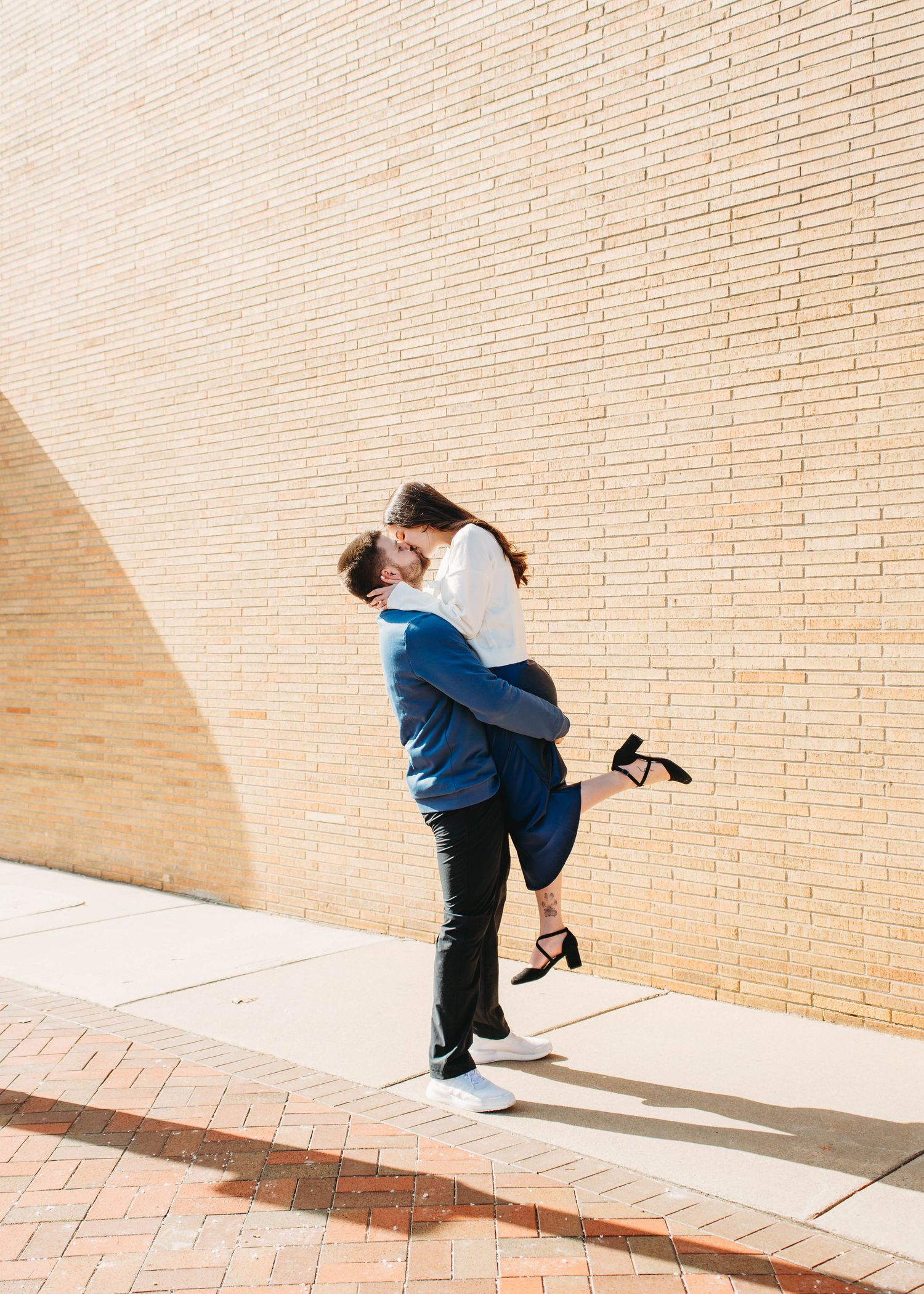 Man lifts and kisses woman in front of a sunlit brick wall outdoors.