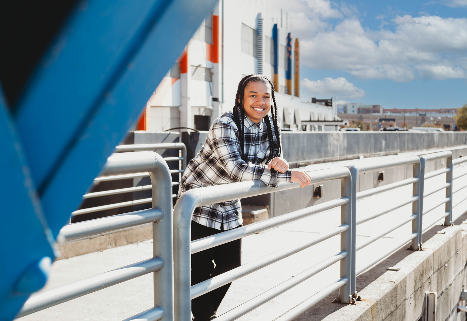 Smiling young woman in plaid shirt leaning on outdoor railing on sunny day.