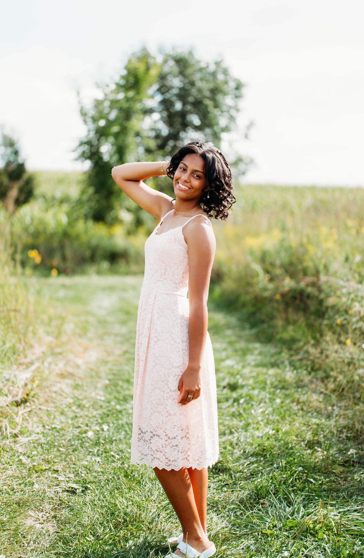 Young woman in pink lace midi dress smiling in green field.