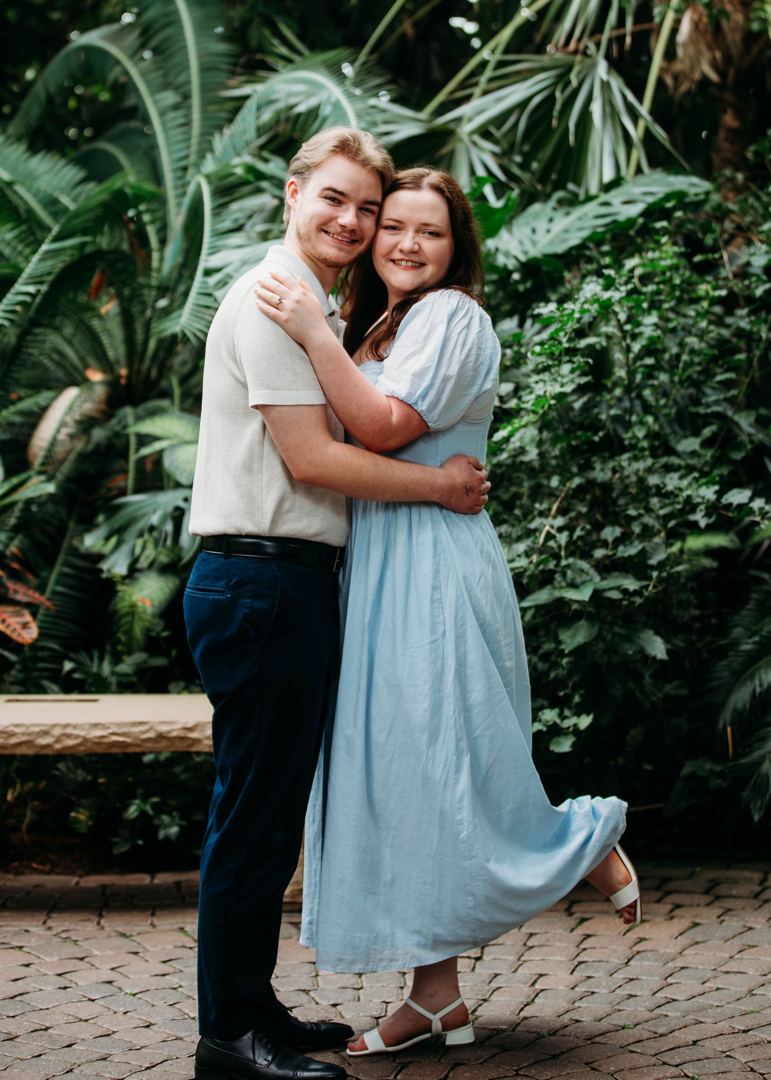 Smiling couple hugging warmly inside a lush tropical greenhouse with palm trees behind them.