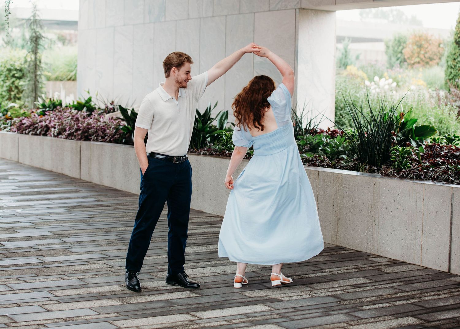 Man spinning woman in a light blue dress on an outdoor stone patio near greenery.