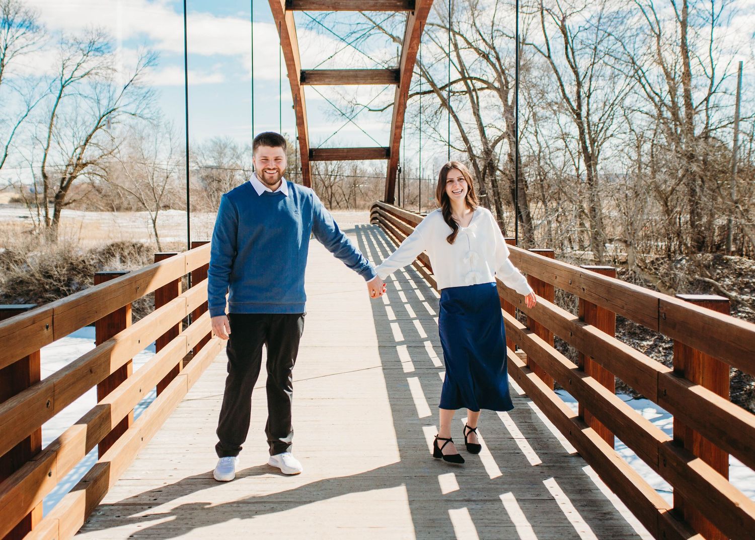 Engaged couple holding hands and smiling on a wooden bridge outdoors on a sunny winter day.