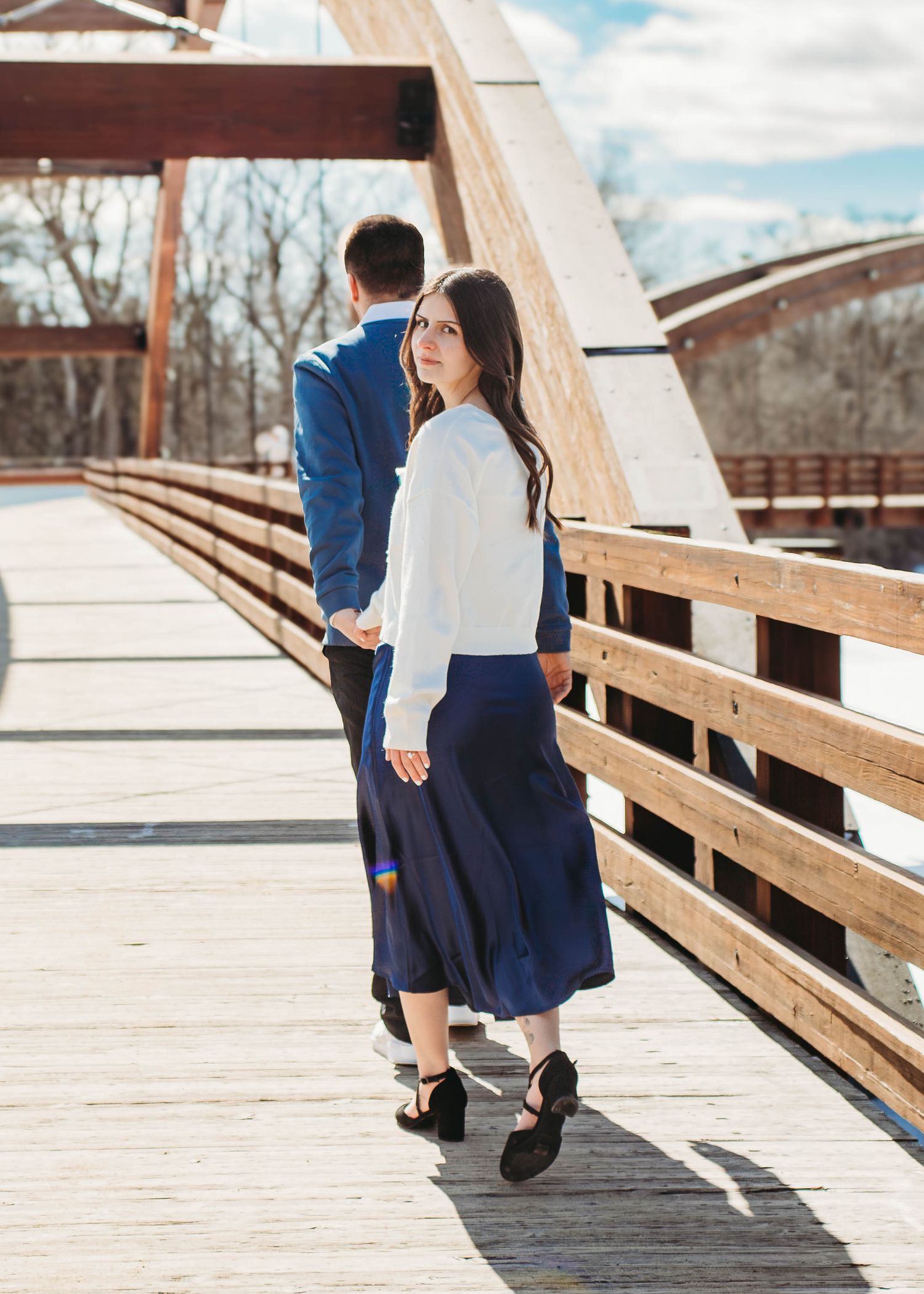 Woman looks back at camera while holding hands with man walking on a sunny bridge.