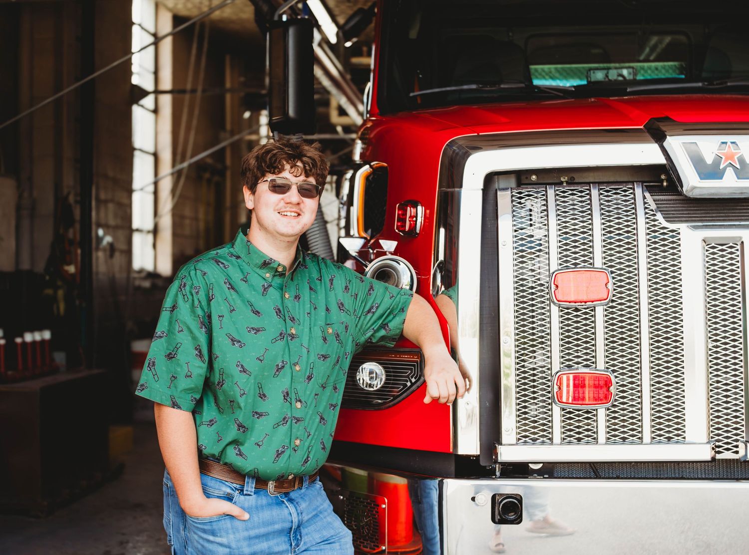 Young man in green shirt smiling while leaning against a red fire truck in a garage.