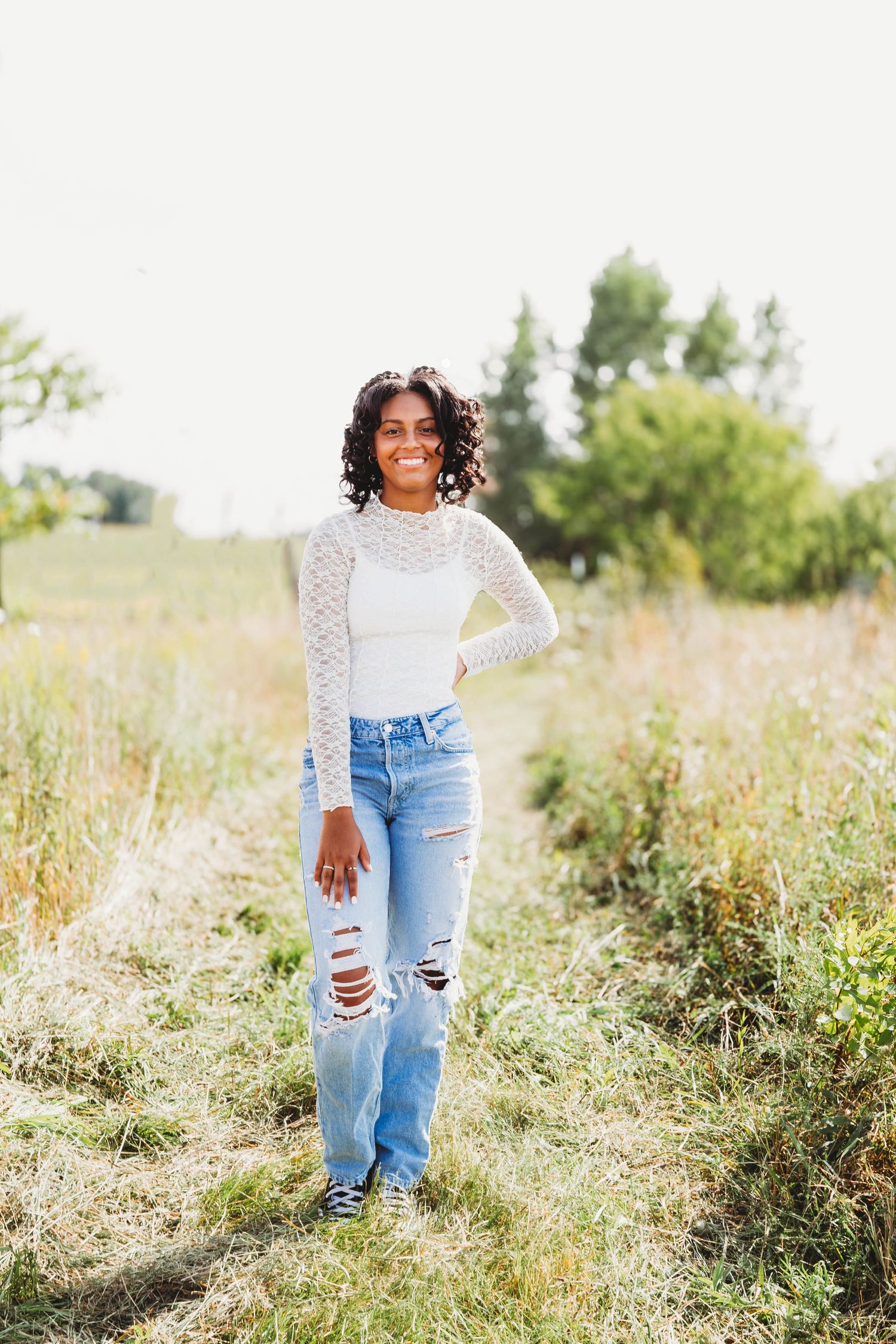 Smiling young woman in white lace top and ripped jeans standing in a sunny meadow field.