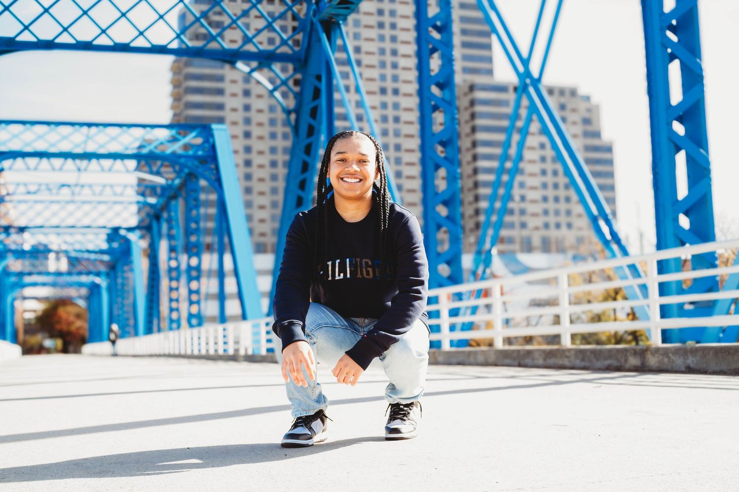 Smiling teen crouching on blue bridge in black hoodie.