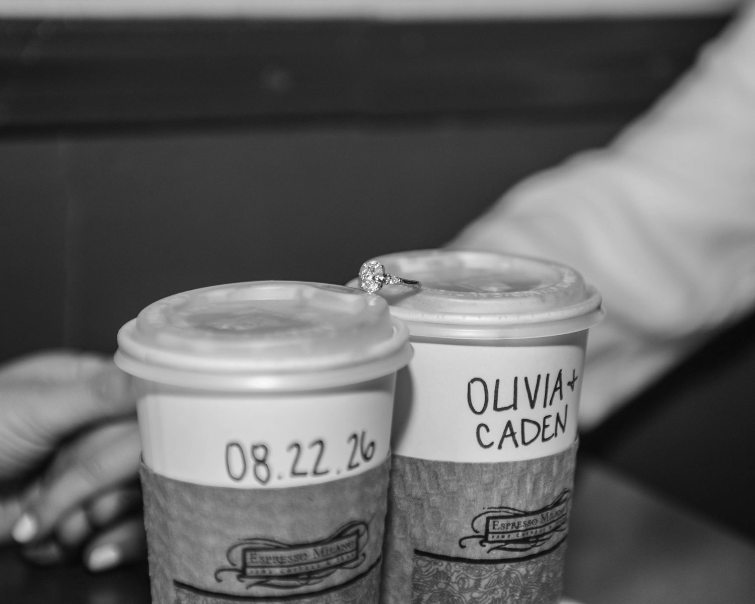 Two coffee cups with engagement date 08.22.26 and names Olivia and Caden written on them with a diamond ring on top.