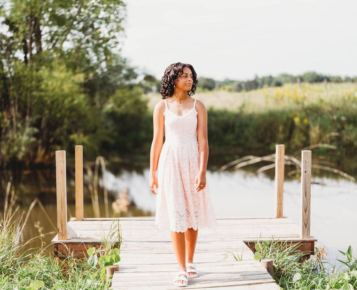 Woman in pink lace dress standing on wooden dock by lake.