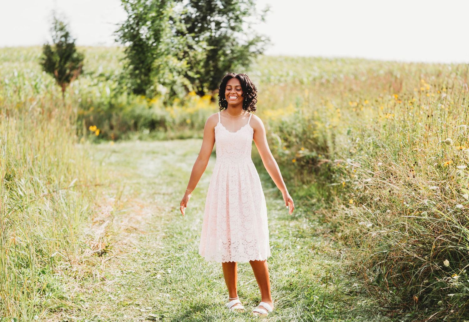Woman in blush lace dress walking on sunny meadow path.
