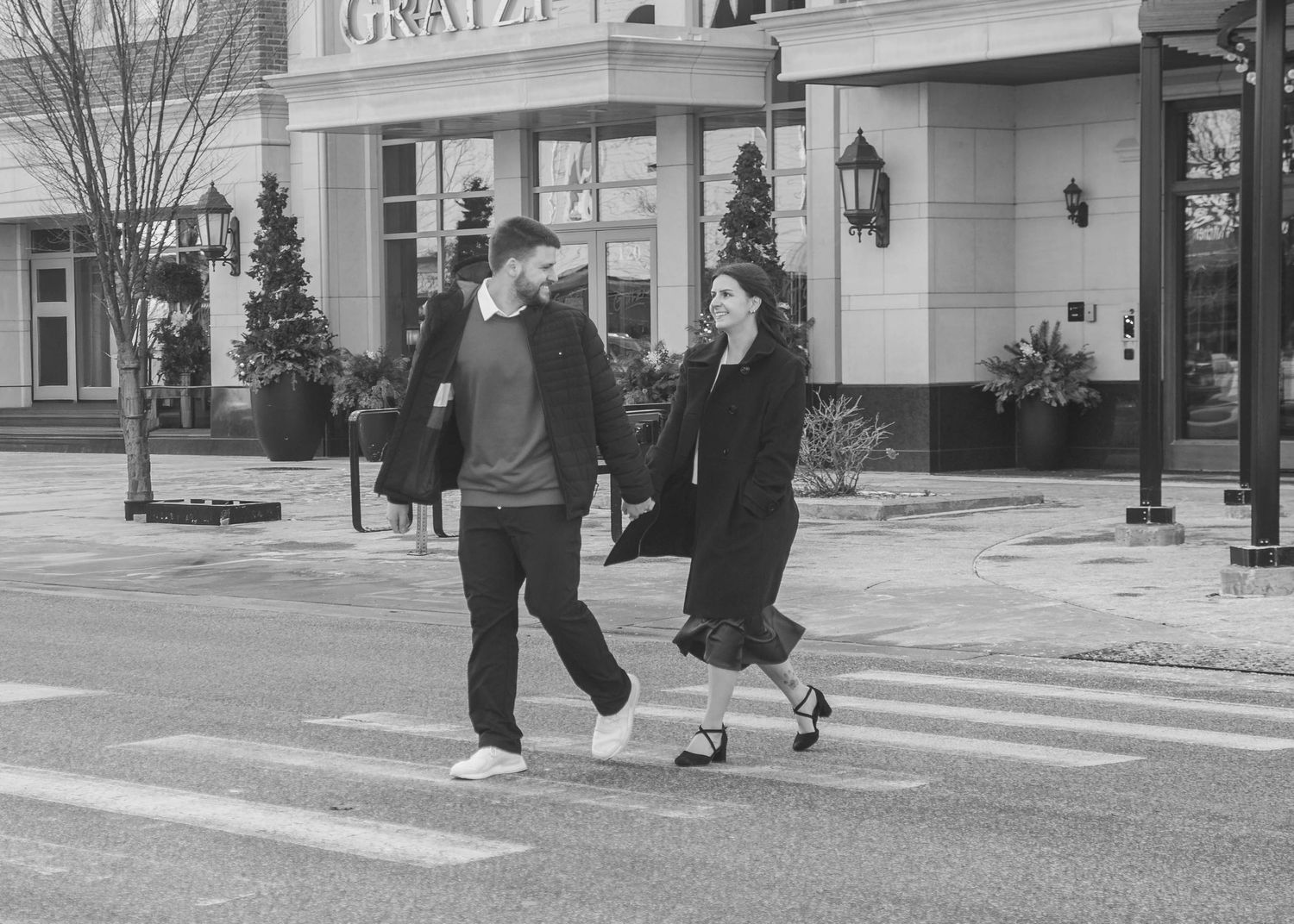 Black and white photo of a couple crossing a street in front of a hotel.