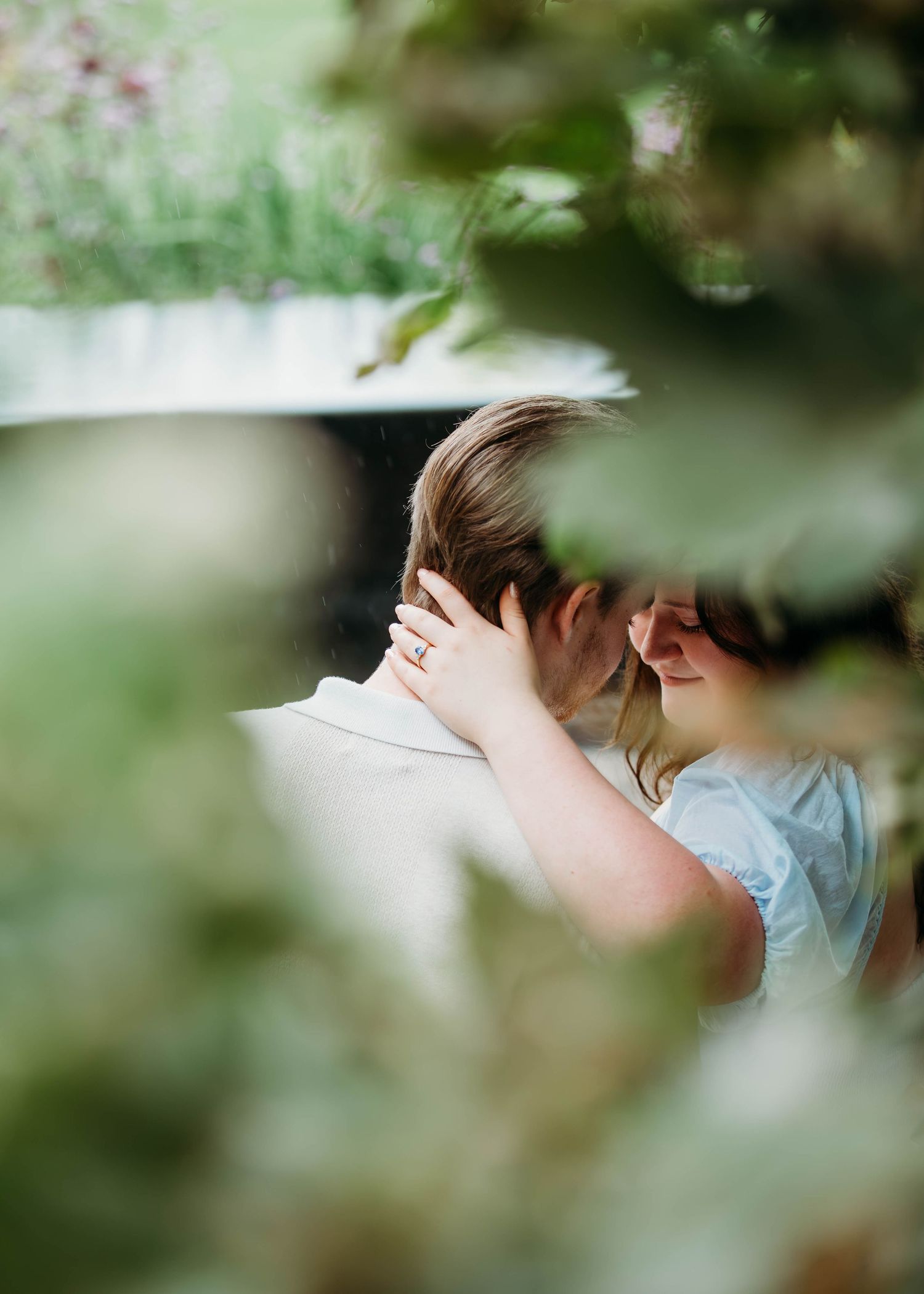 Couple sharing intimate moment framed by blurred green leaves during outdoor engagement session.