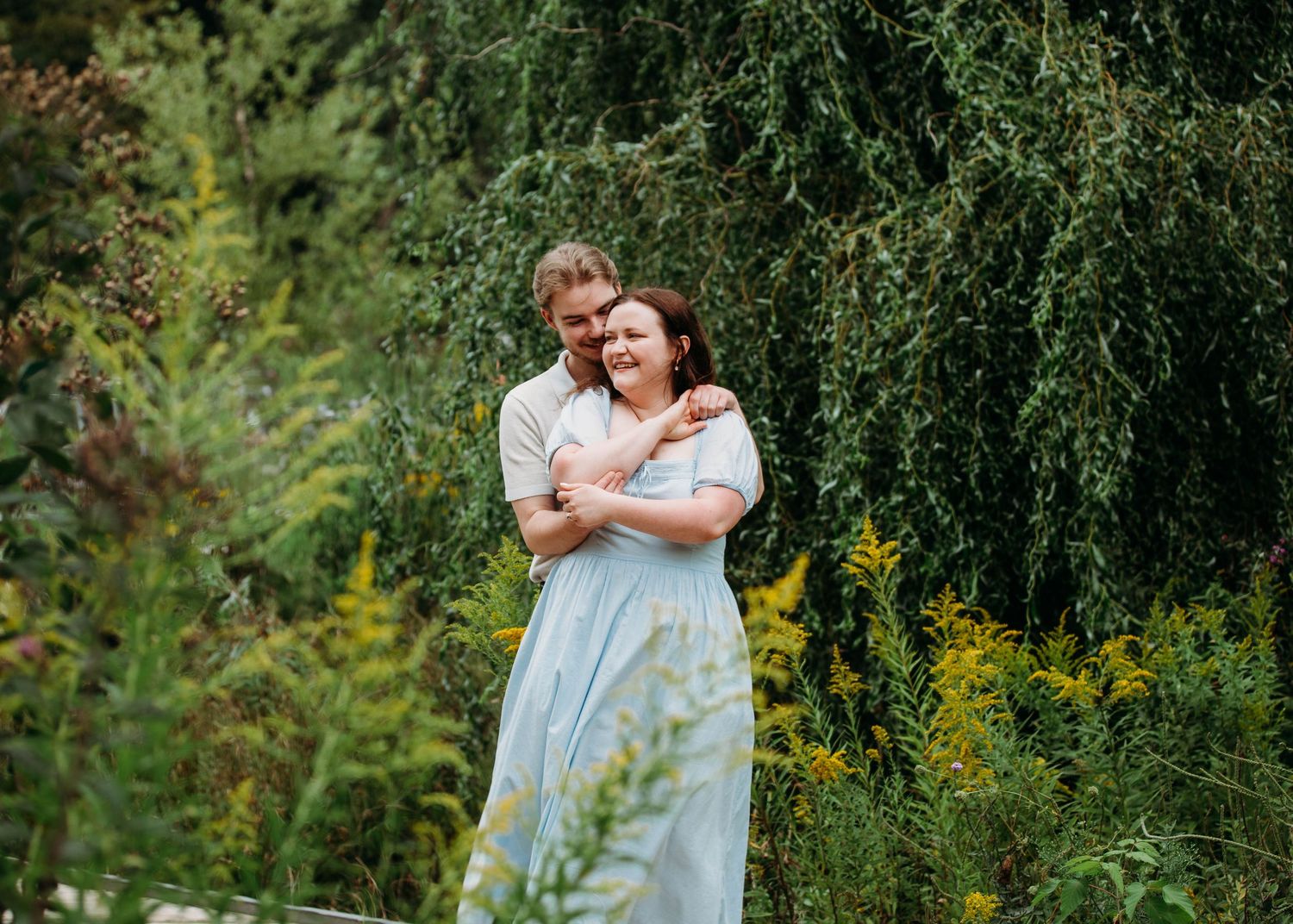 Happy couple embracing and laughing together surrounded by green weeping willow and wildflowers.