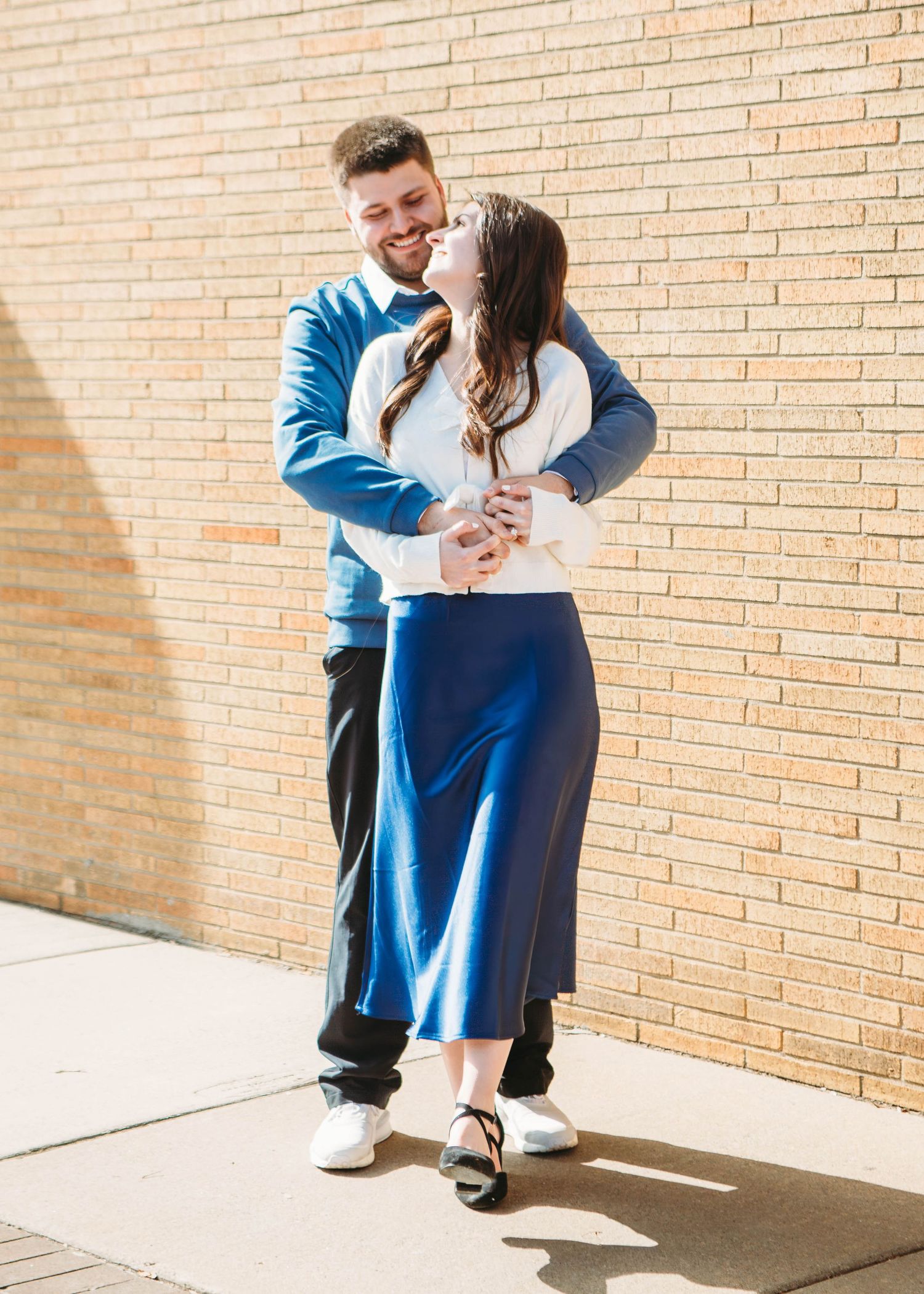 Couple embracing and smiling against a sunny brick wall, woman in blue midi skirt and white top.