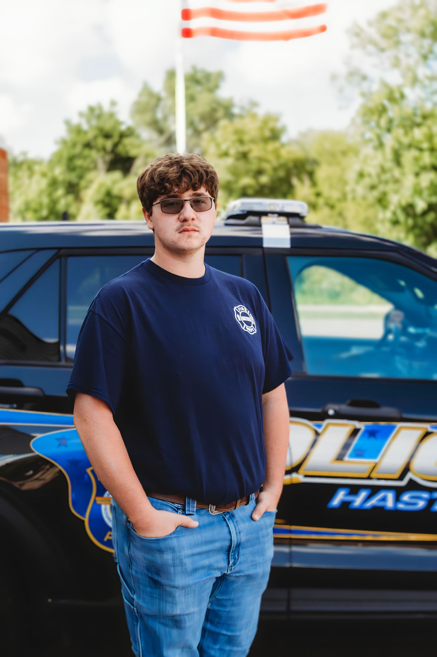 Young man in navy shirt and sunglasses standing confidently in front of a police vehicle.