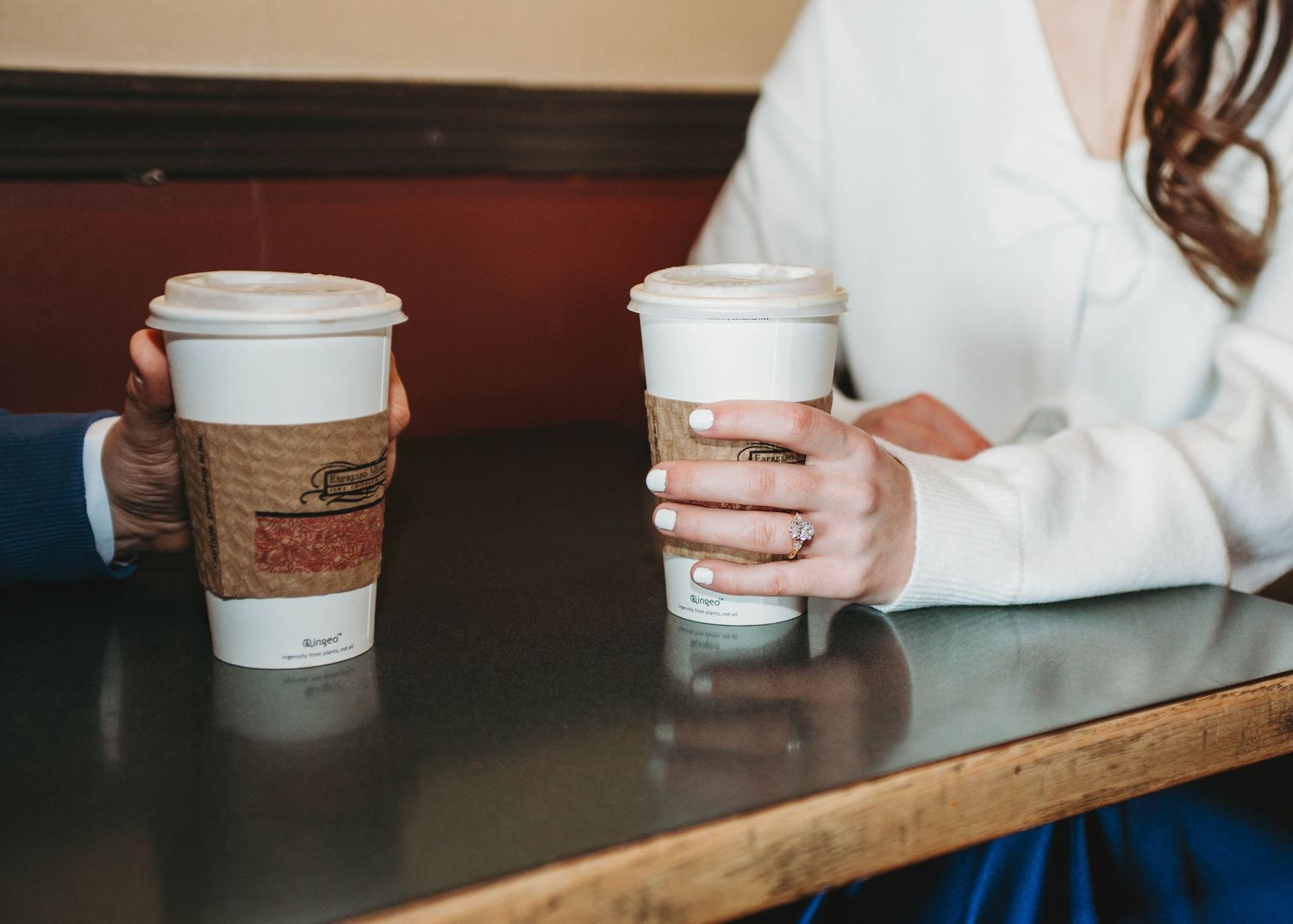 Couple holding matching coffee cups with brown sleeves at a cafe table during engagement session.
