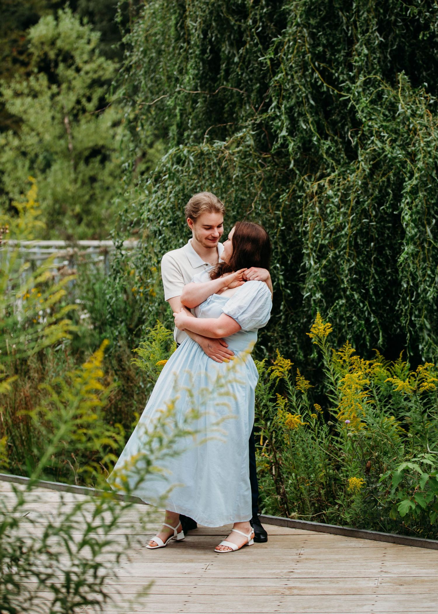 Joyful couple dancing together on a wooden deck surrounded by weeping willow and yellow flowers.