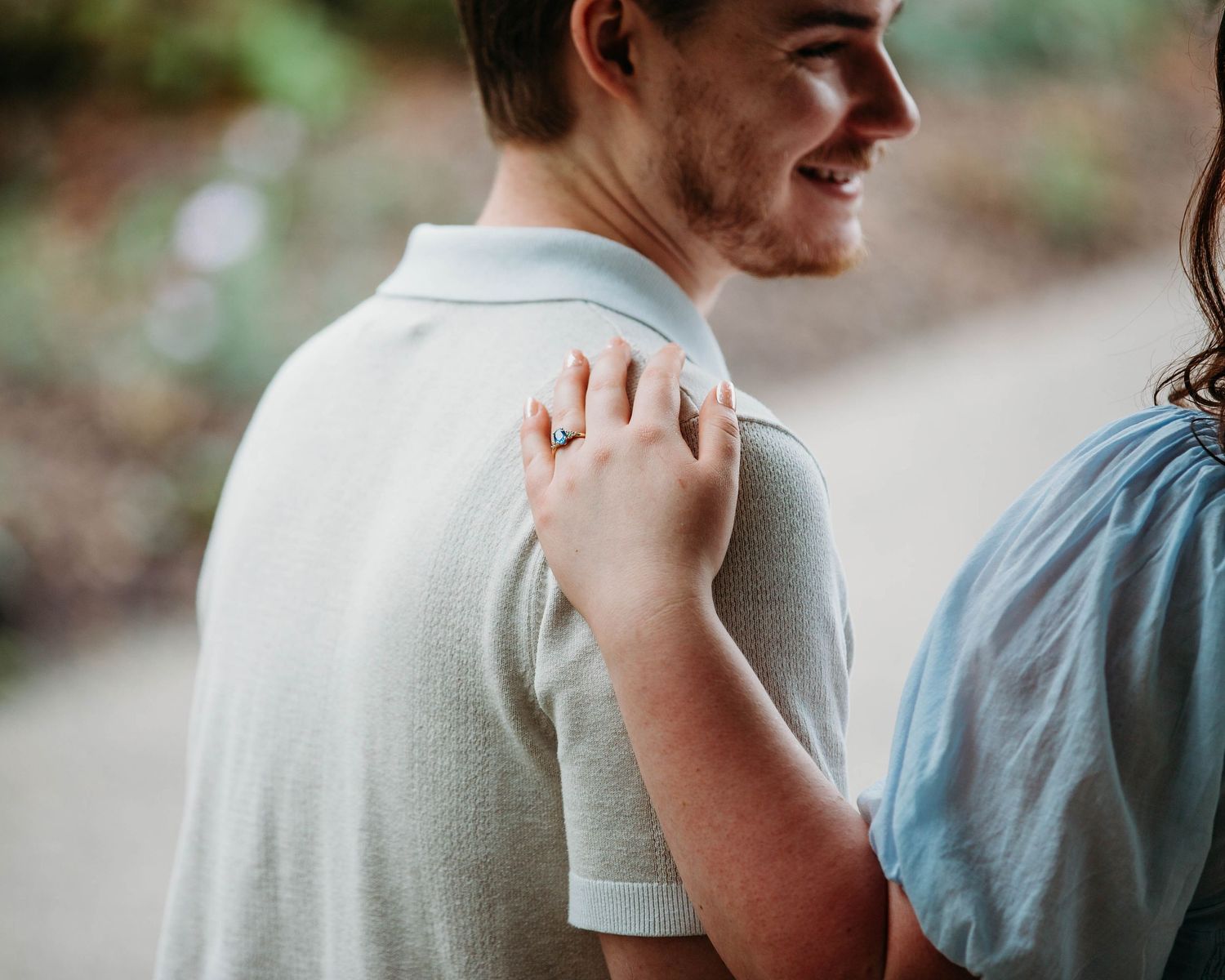 Close-up of woman's hand with engagement ring resting on man's shoulder in light grey polo.