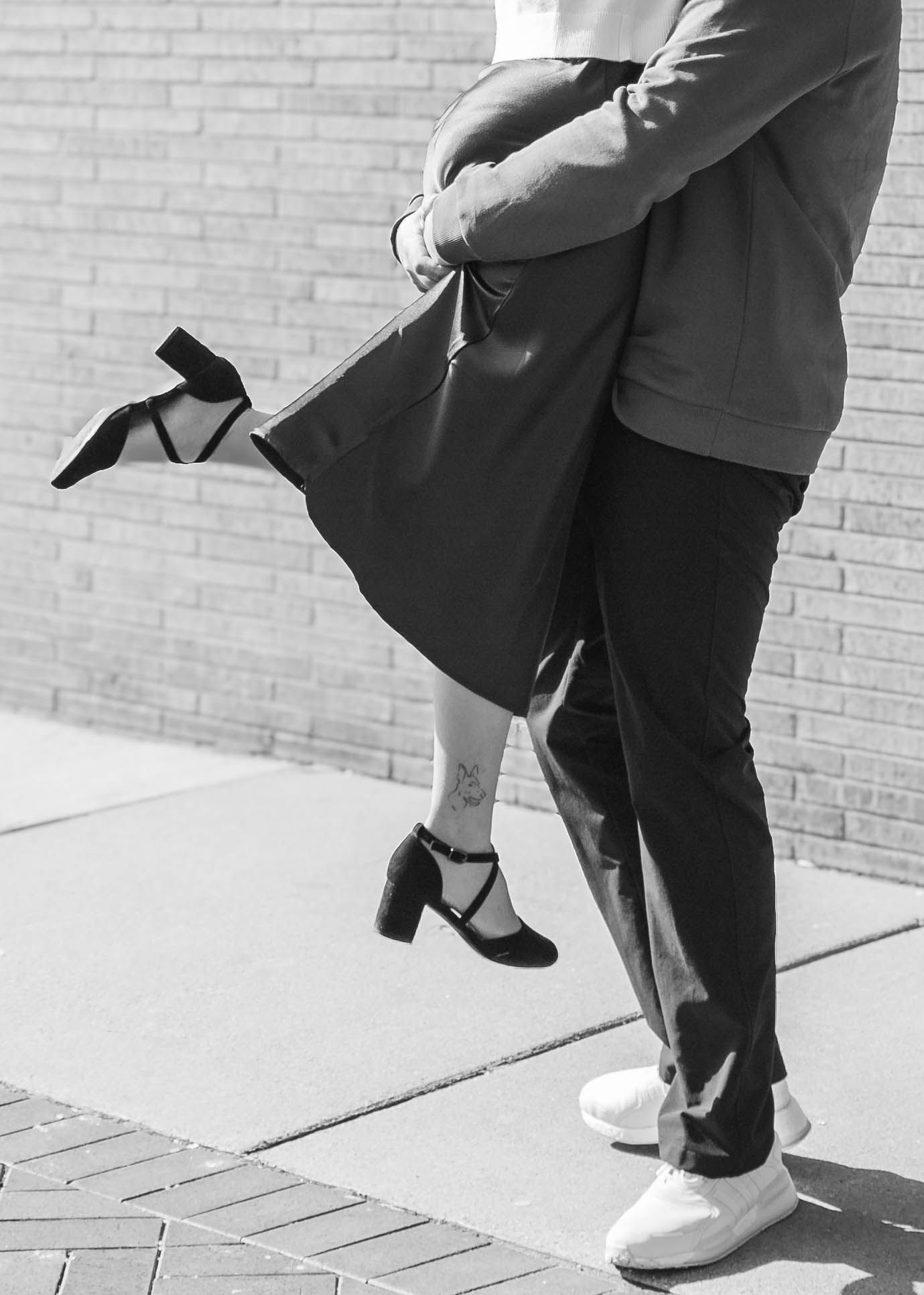 Black and white close-up of couple embracing, woman lifted in black heels against brick wall.