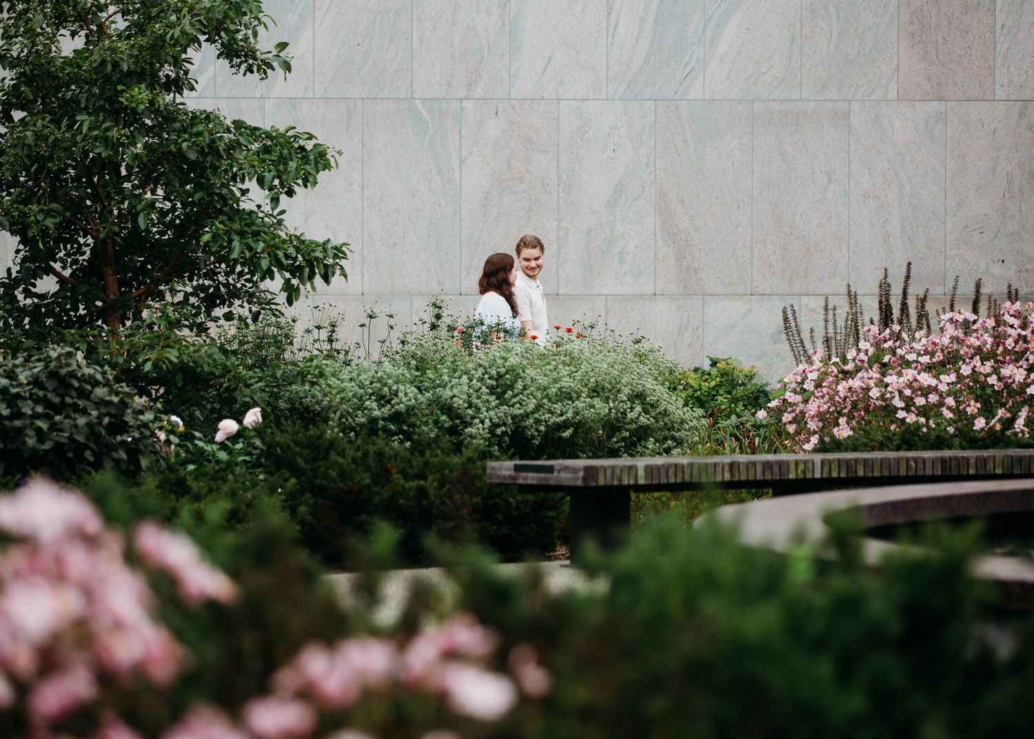 Engaged couple walking through a blooming garden with pink roses near a stone wall.