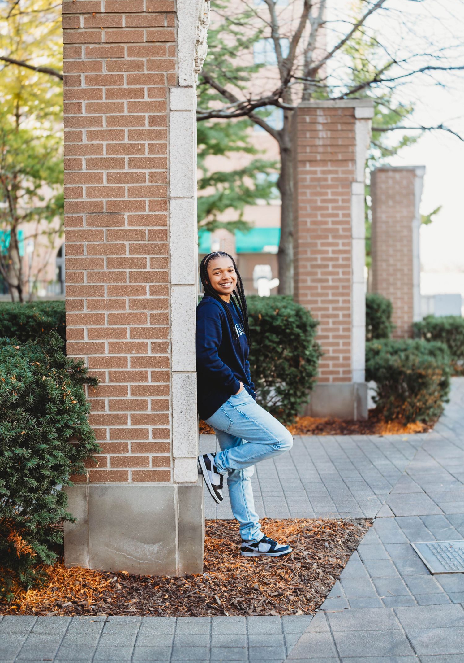 Teen leaning on brick pillar in navy hoodie and jeans.