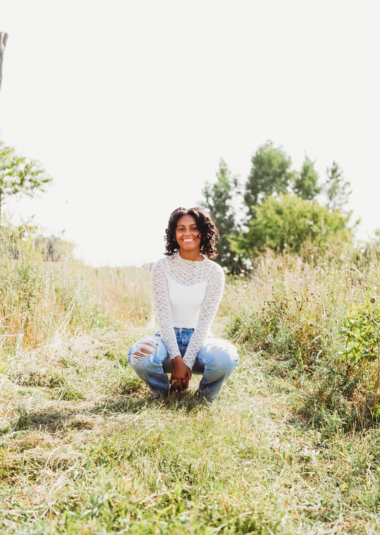 Young woman in white lace top and distressed jeans sitting cross-legged in a grassy meadow.