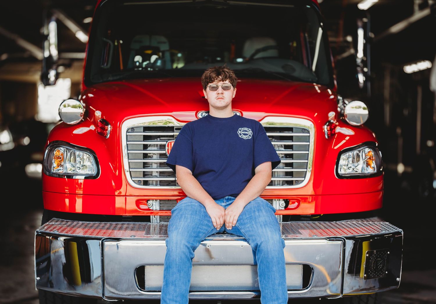 Teen boy in navy shirt sitting on the front bumper of a red Freightliner fire truck.