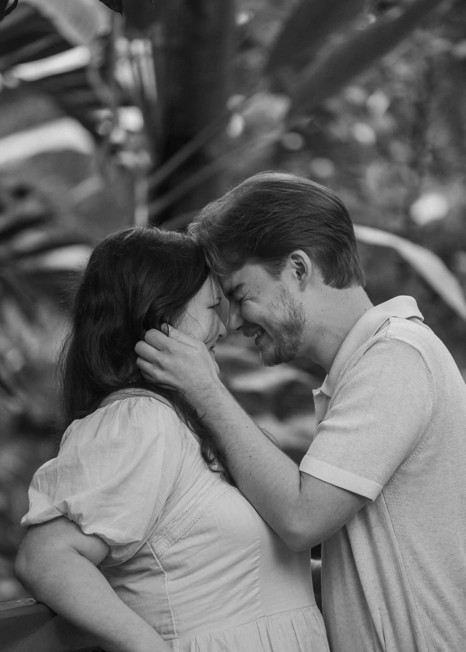 Black and white photo of couple laughing and touching foreheads in a tropical garden setting.