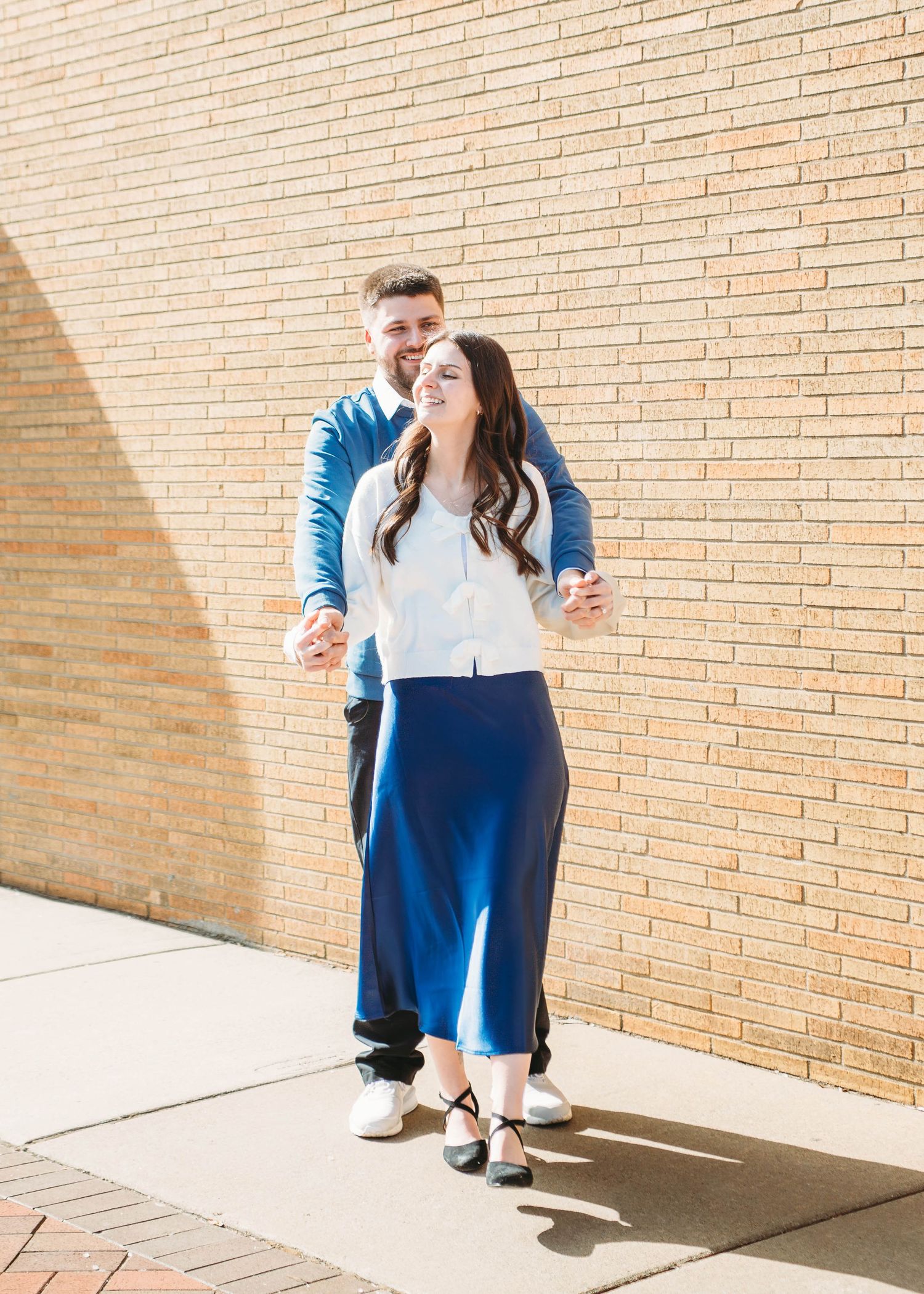 Couple laughing and walking together along a sunny brick wall outdoors.
