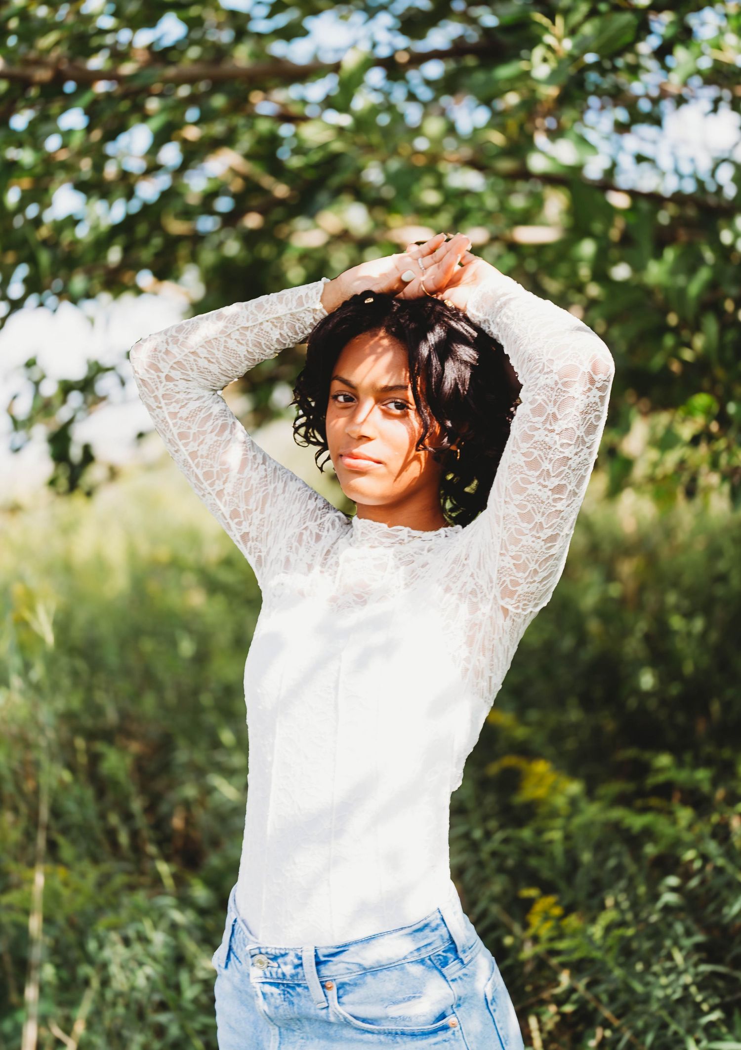Young woman in white lace top raising arms overhead while standing beneath a leafy tree outdoors.