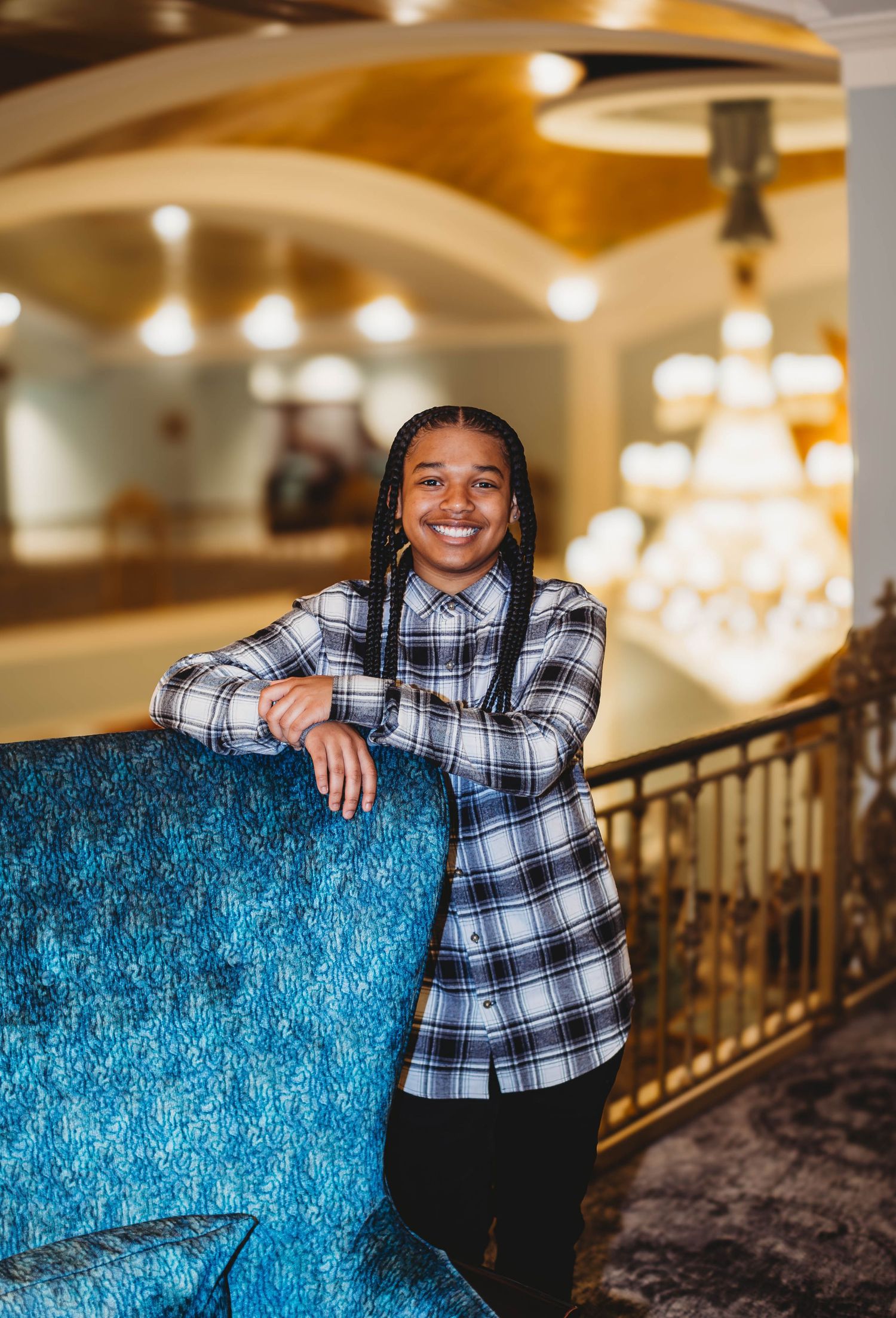 Happy young woman with braids in plaid shirt leaning on blue couch in elegant ballroom.