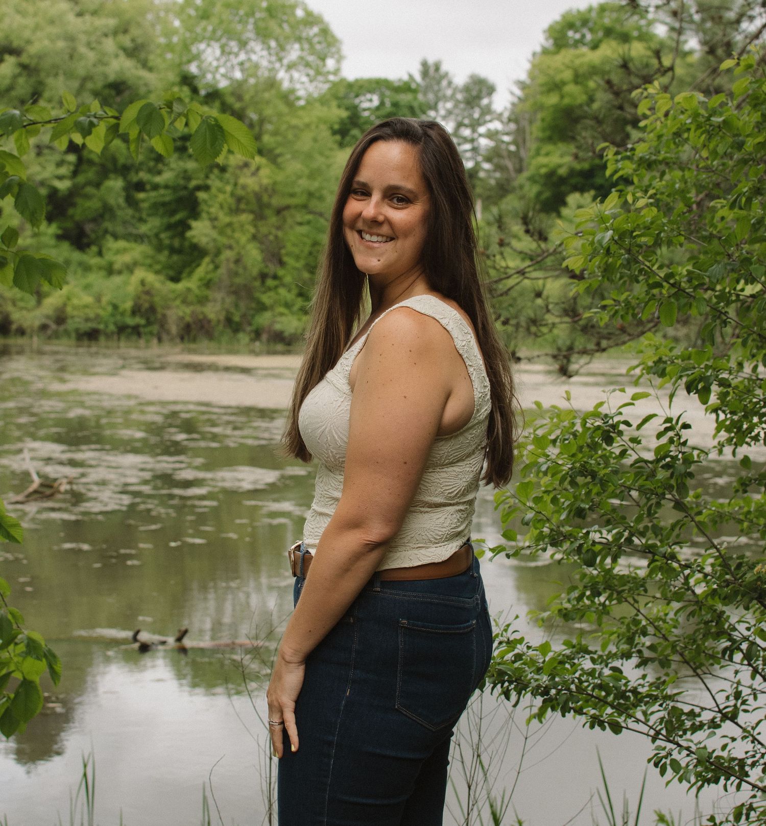 Woman in white top and jeans posing by a serene pond surrounded by lush green trees.