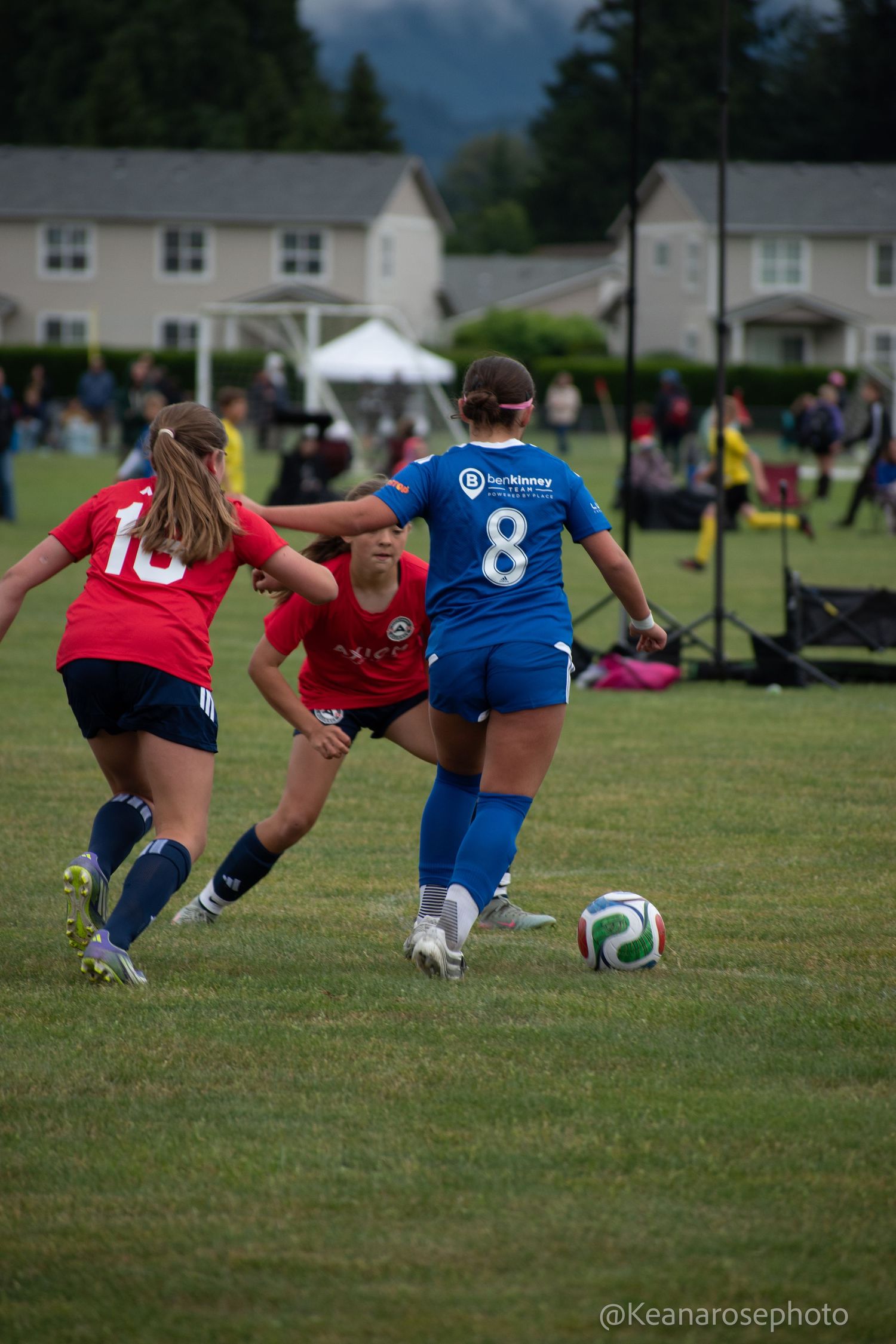 Female soccer players in red and blue uniforms compete for possession of the ball during outdoor match.