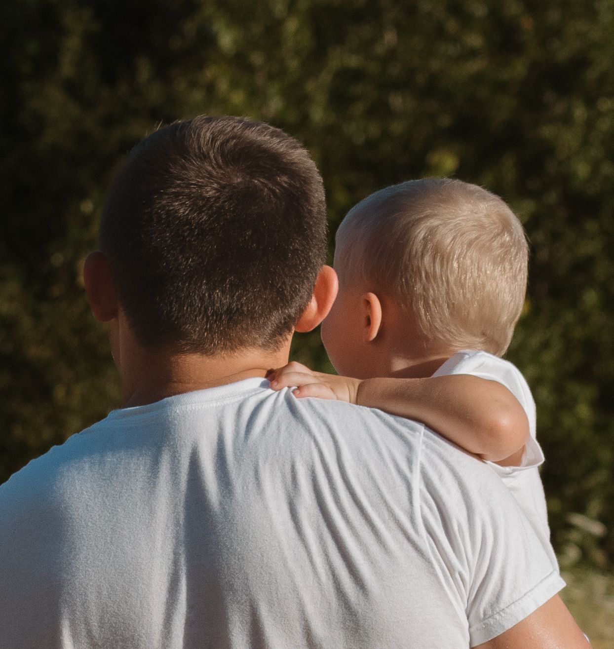 Father in white t-shirt holding baby on shoulder with green trees in background.