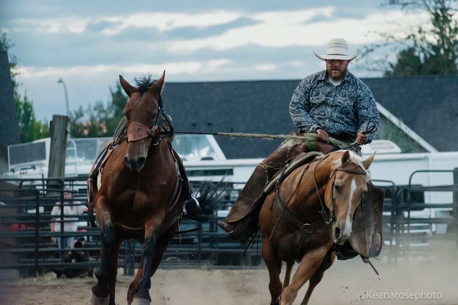 Cowboy on horseback leads a loose brown horse through a dusty rodeo arena.