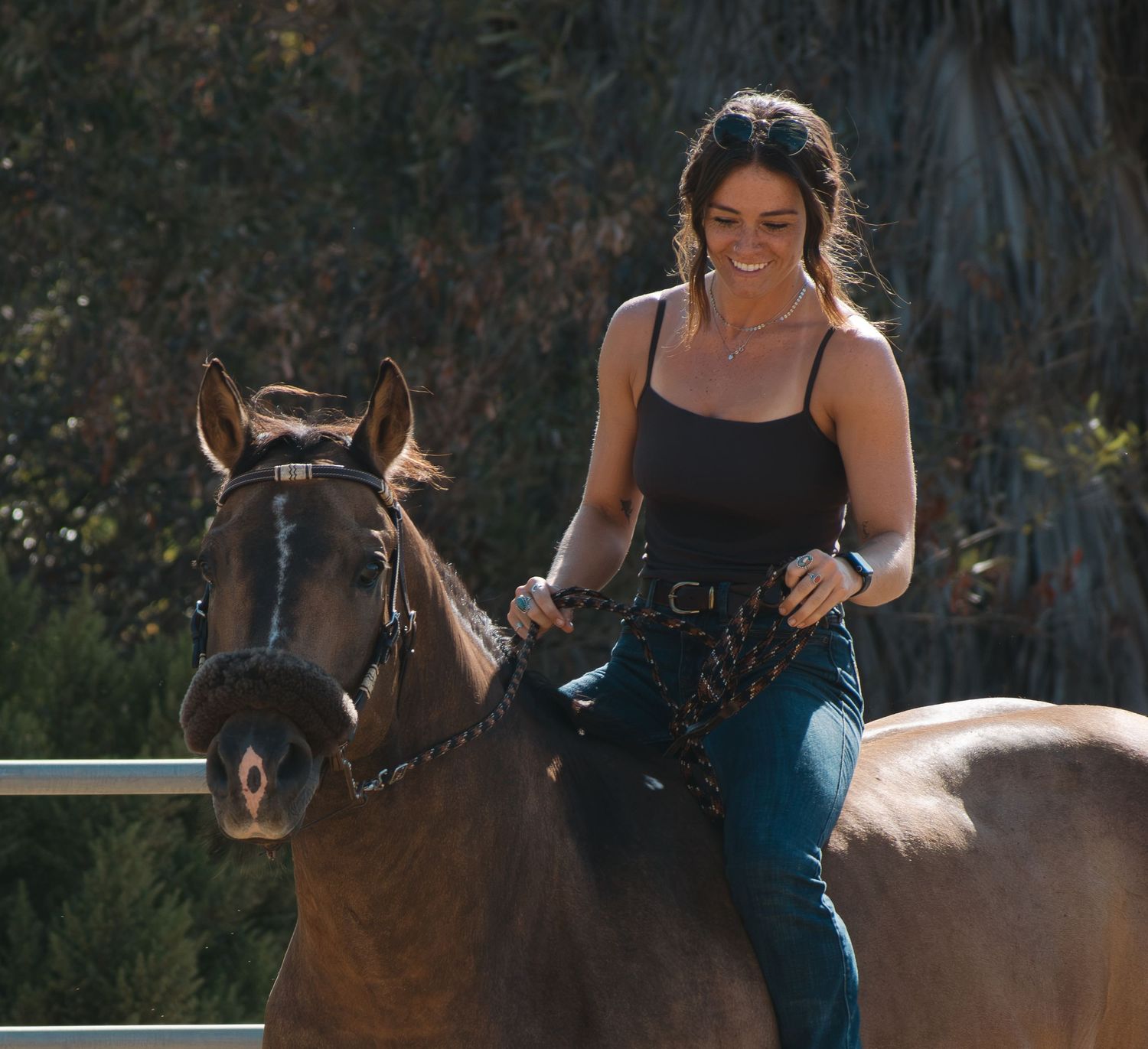 Smiling woman in black tank top riding a brown horse outdoors with trees in the background.