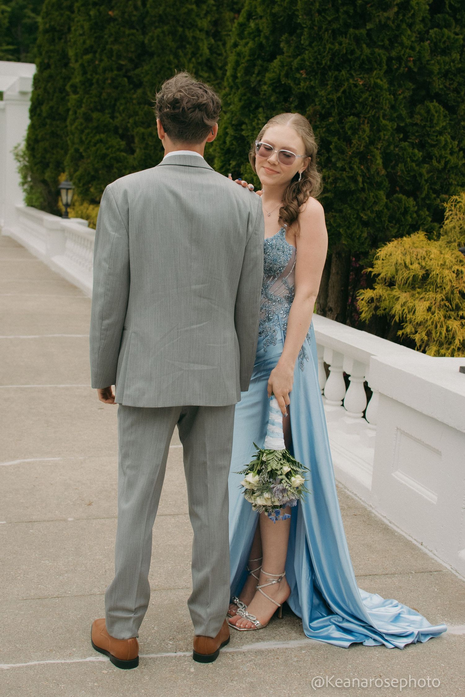 A couple in prom attire walk together, woman in blue gown holding bouquet, man in gray suit, on terrace.