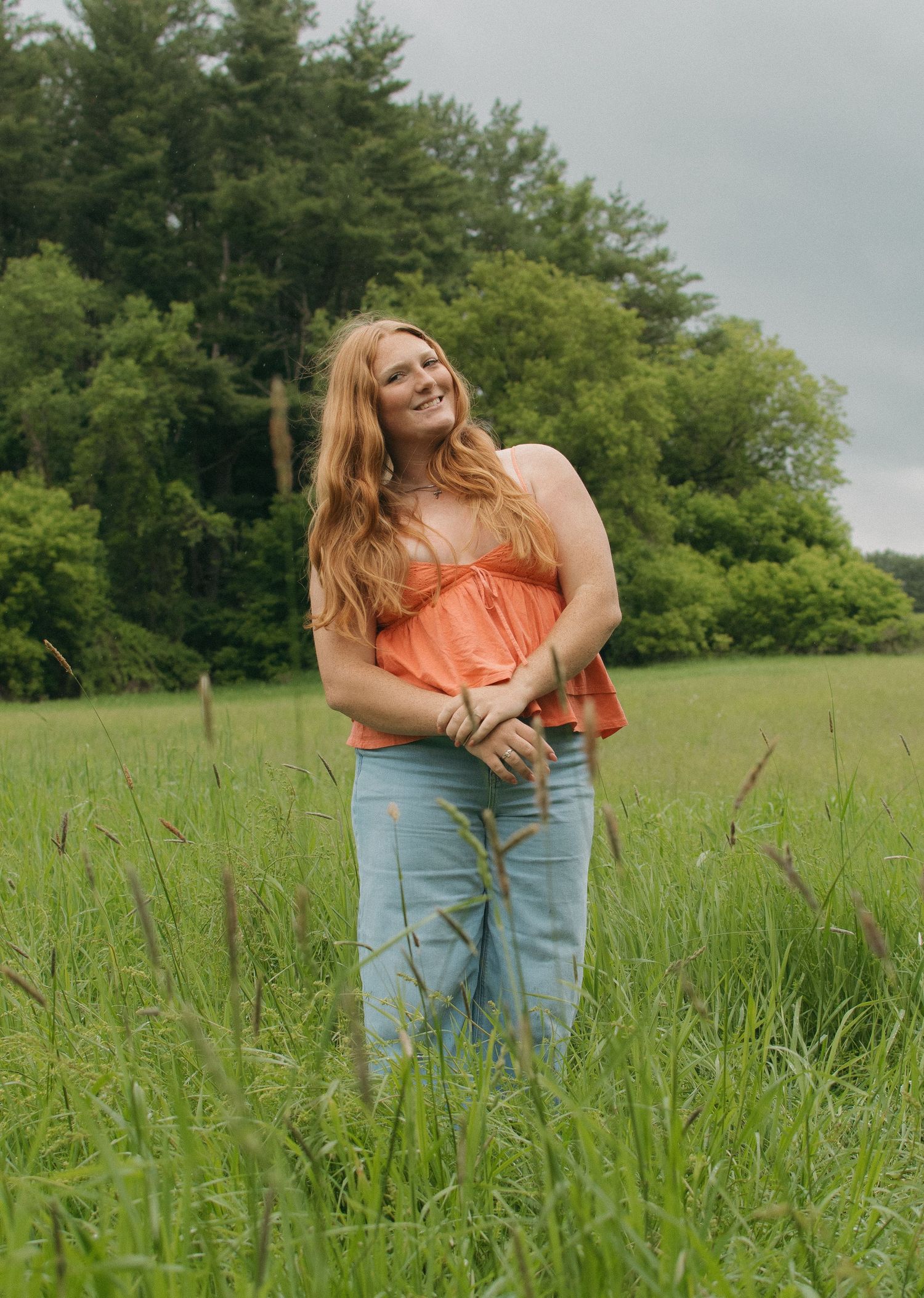 Young woman with long red hair in orange top and jeans smiling in a lush green meadow with trees behind.