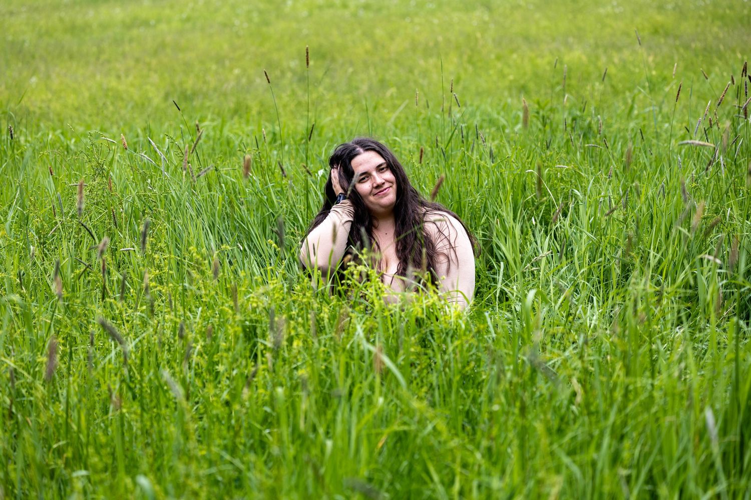 Young woman with long dark hair sitting peacefully in a lush green meadow, looking up and smiling.
