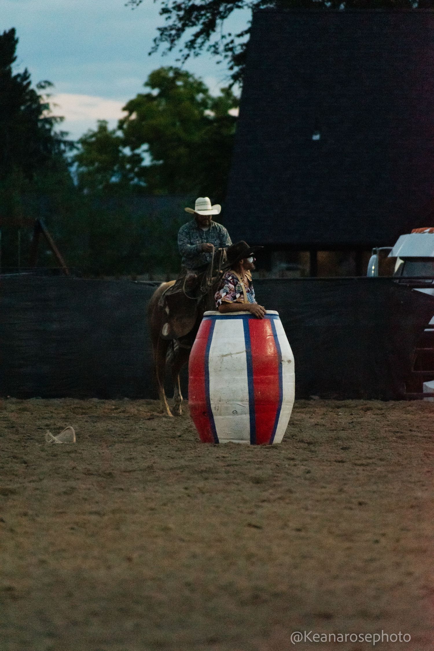 Rodeo clown leans on a patriotic red white and blue barrel as a cowboy on horseback watches.