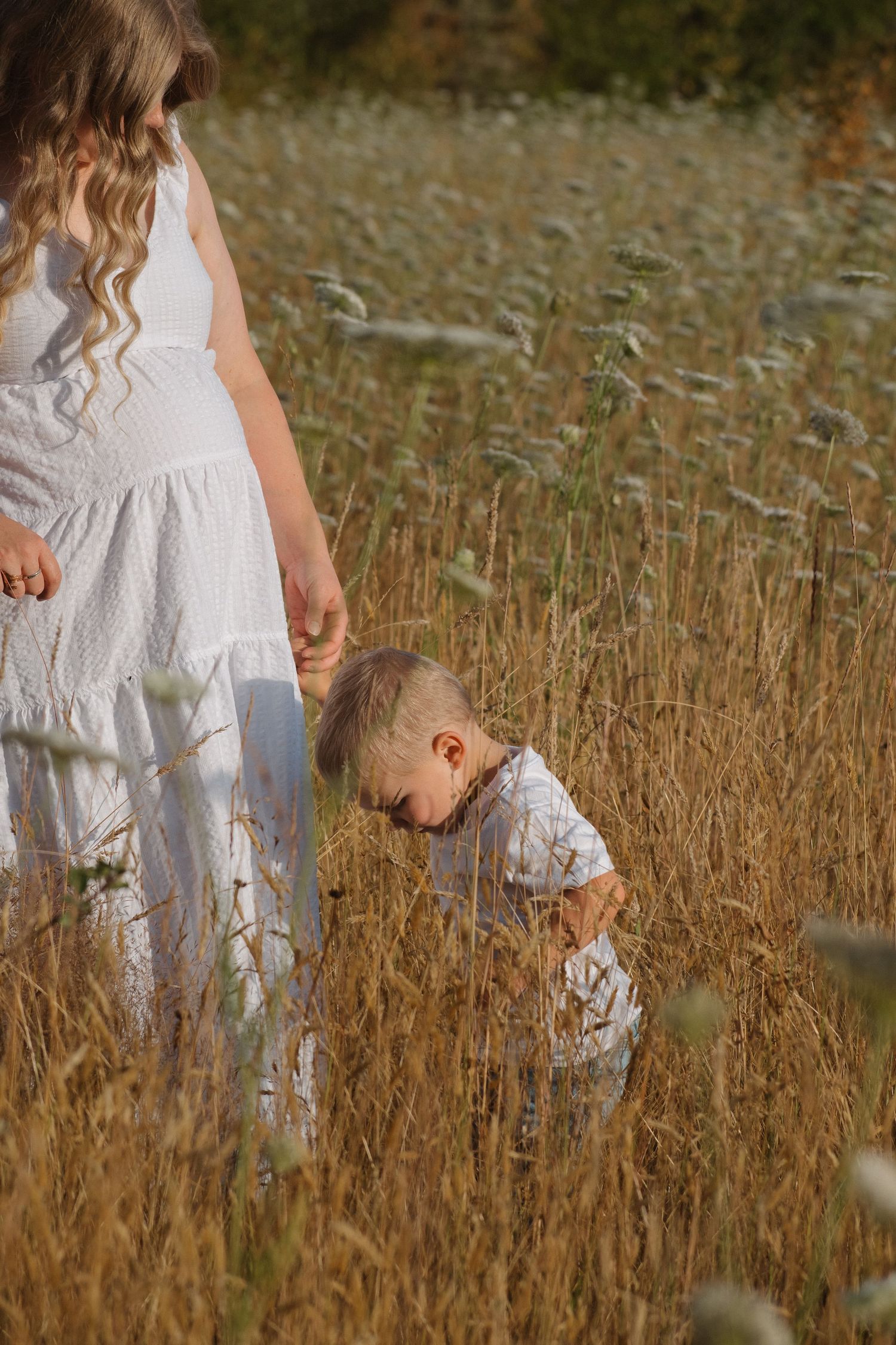 Pregnant woman in white dress holding hands with young boy exploring tall grass field.