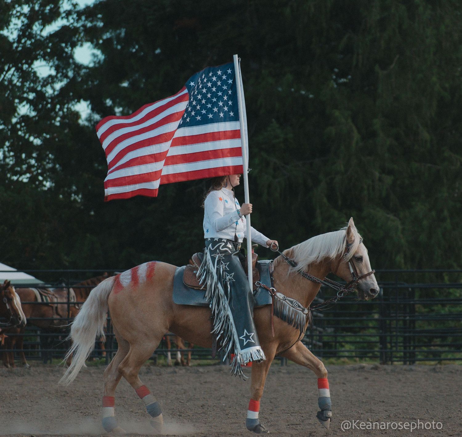 Cowboy rides palomino horse carrying large American flag in dusty outdoor rodeo arena.
