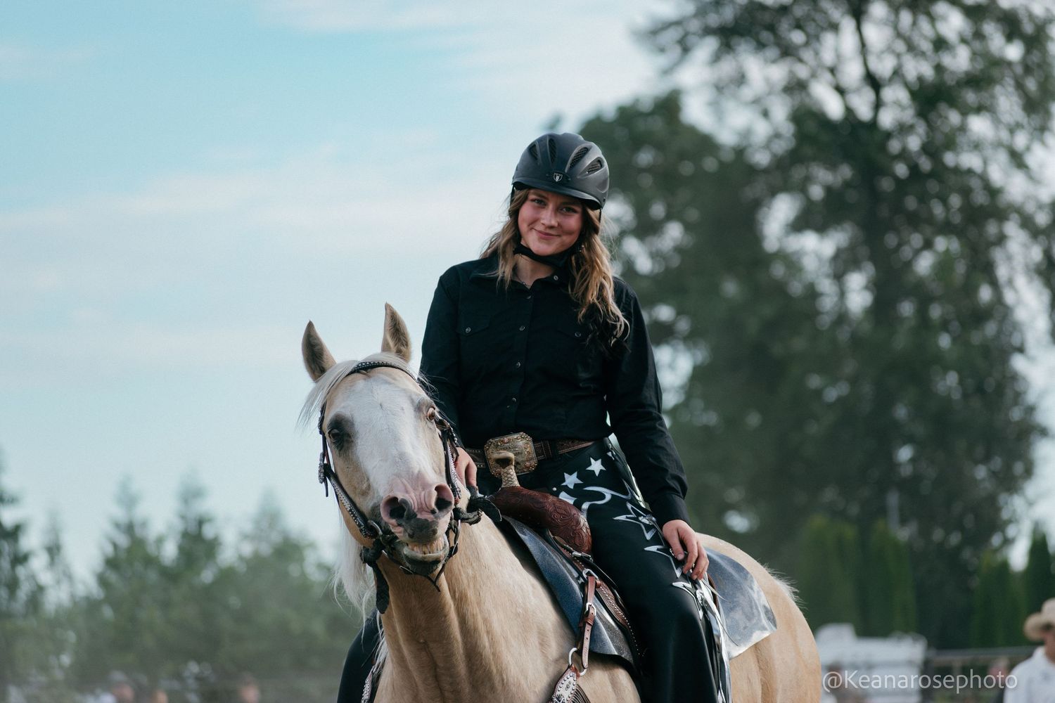 Smiling female rider in black helmet rides palomino horse in outdoor equestrian arena.