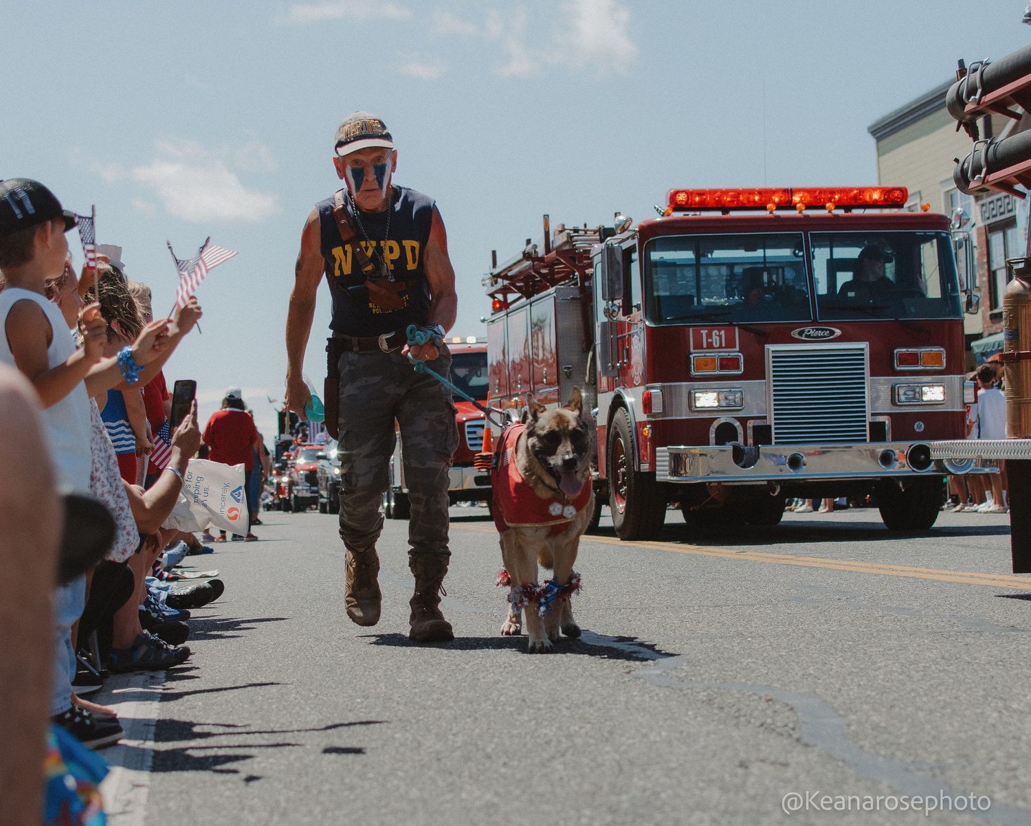 Veteran walks dog in Fourth of July parade with fire truck behind them as crowds wave American flags.